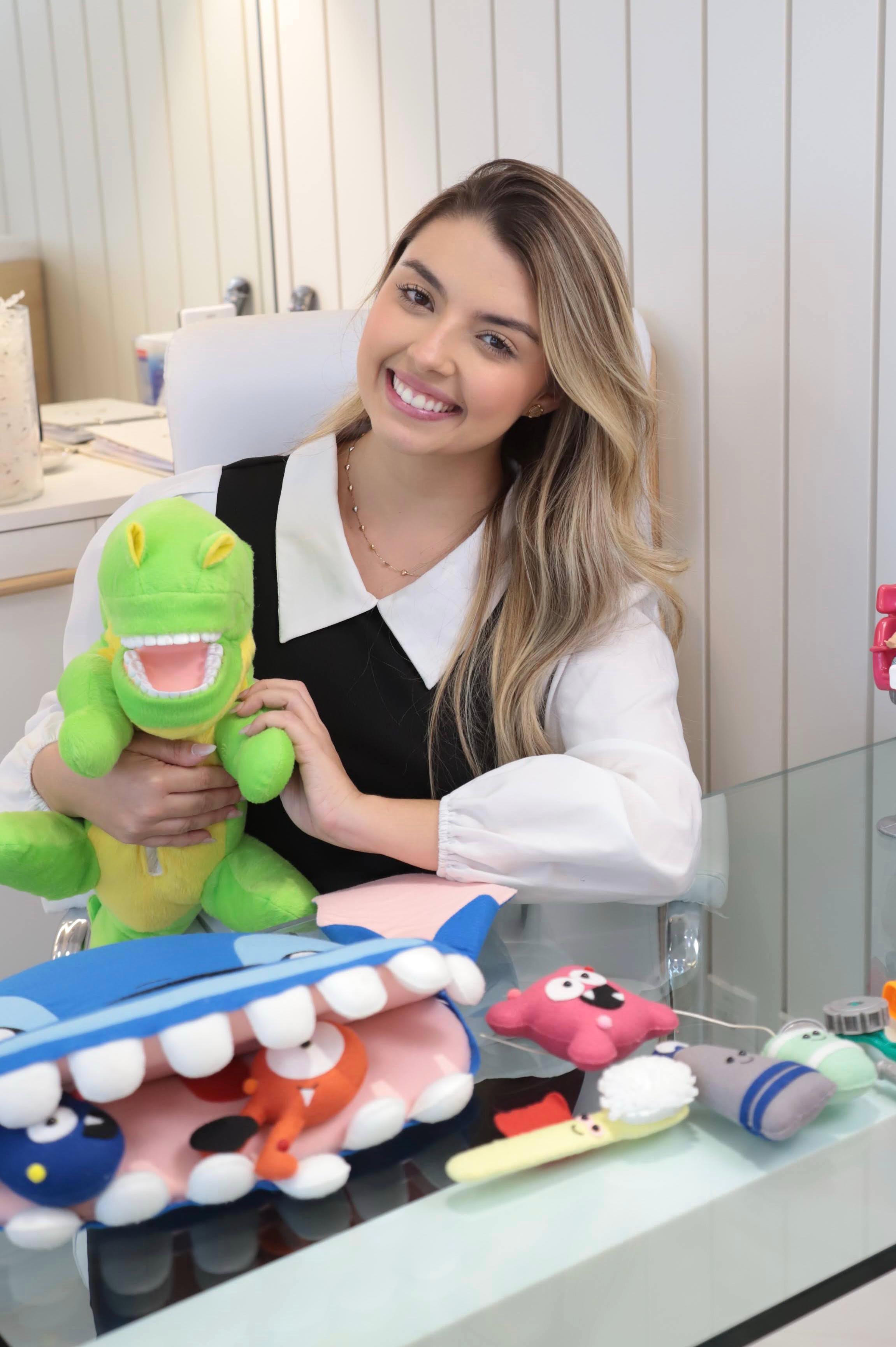 Dra. Thirza Nunes, odontopediatra em São Luís, sorrindo em seu consultório enquanto segura um dinossauro de pelúcia usado nas consultas infantis